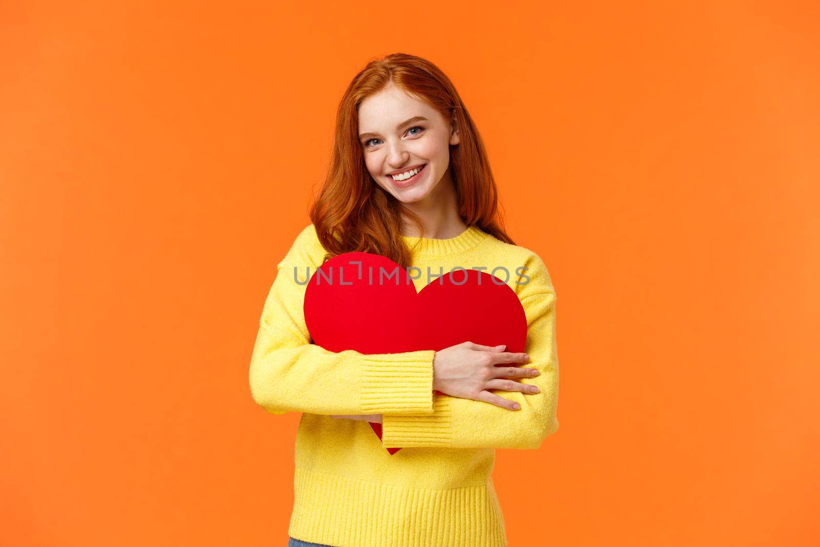 Valentines day, romance or relationship concept. Tender, lovely redhead girl waiting for her loved one, receive valentine card, hugging large heart sign and smiling happy, standing orange background.