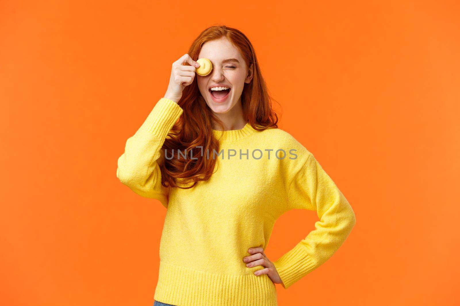 Happy and carefree smiling, laughing lovely redhead girl grinning, holding tasty macaron near eye, fooling around, having fun, eating delicious dessert at favorite cafe, orange background.