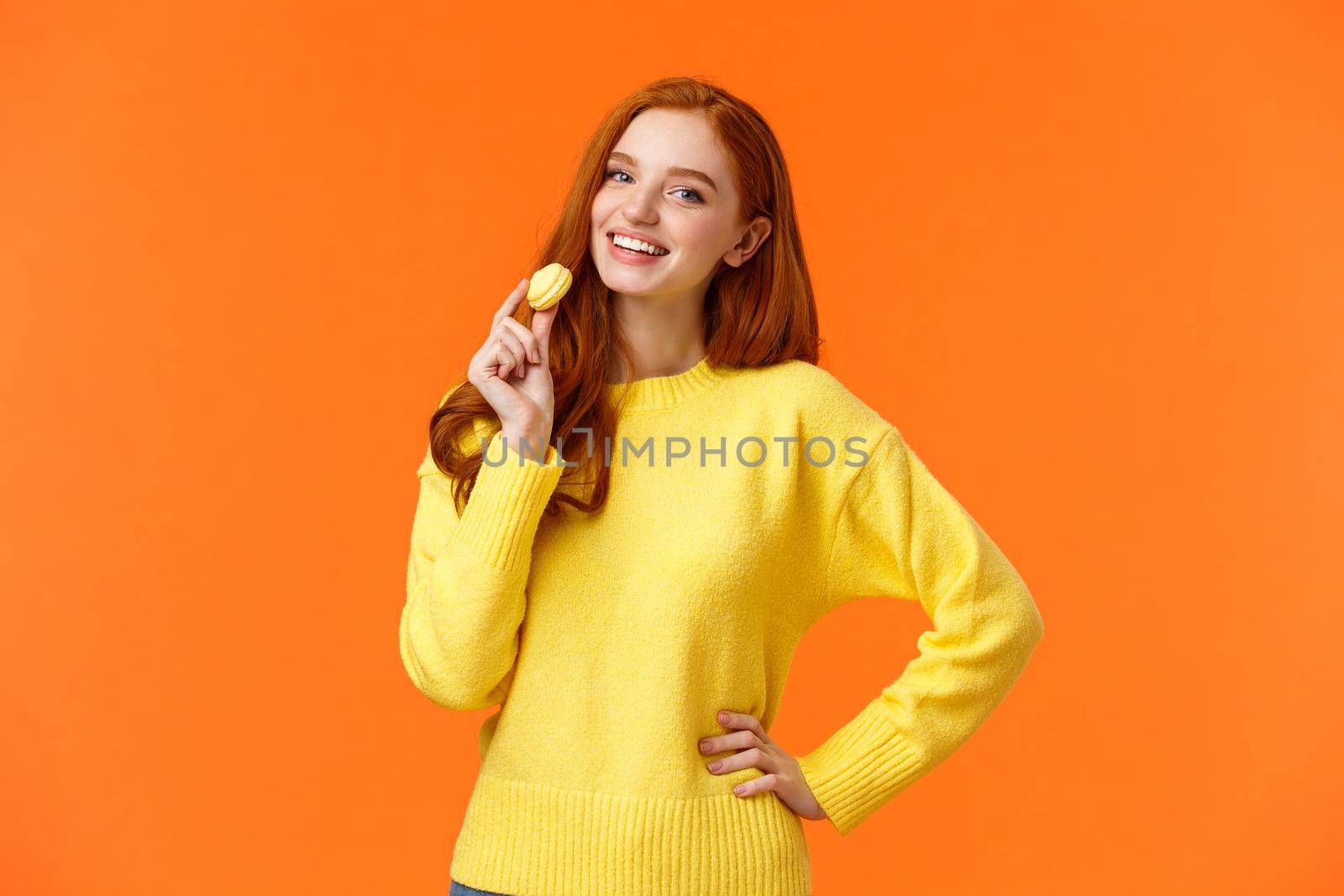 Waist-up portrait lovely redhead girl holding tasty yellow macaron near mouth and smiling joyfully, like eating sweets, buy dessert for romantic valentines date, posing cheerful orange background.