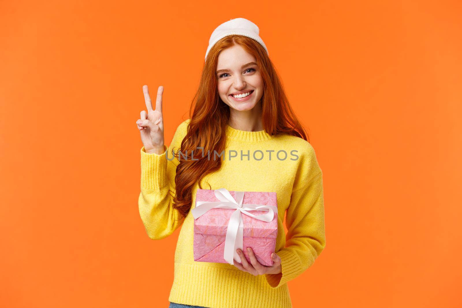 Celebration, holidays and presents concept. Cute cheerful redhead female in winter hat, sweater, holding pink present, showing peace sign as receive gift and smiling delighted, orange background.