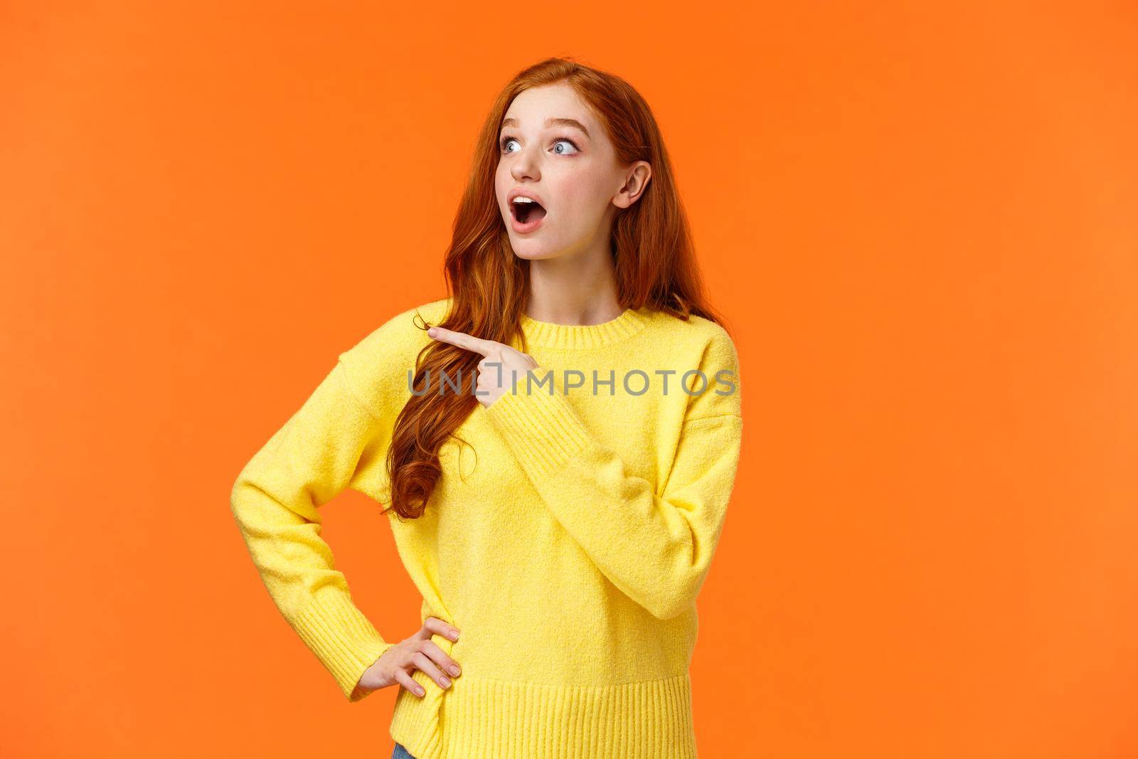 Impressed redhead woman checking-out awesome product sale, stare and pointing left, popping eyes on promo and drop jaw from amazement, surprised, telling about cool advertisiment, orange background.