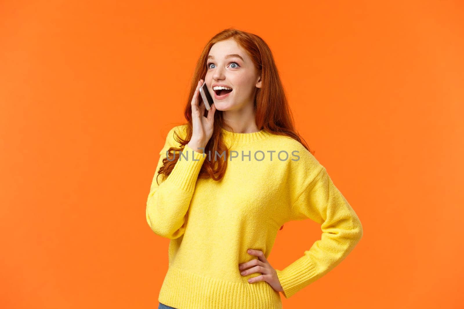 Amused and surprised smiling redhead girl widen eyes and look impressed as hear good news from friend during talking on phone, holding smartphone near ear, have conversation, orange background by Benzoix