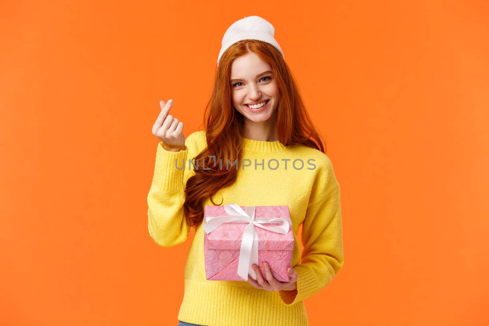 Lovely redhead girlfriend got gift from her girl, showing korean heart gesture and smiling silly, holding pink present box, wearing winter beanie, standing over orange background happy.