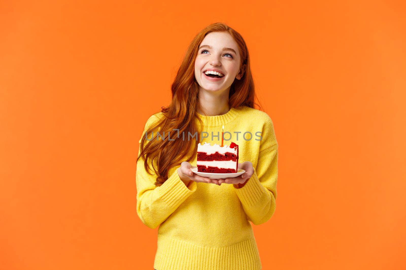 Dreamy happy girl celebrating birthday, having fun, laughing and gazing upper left corner thoughtful, making wish as blowing out candle on delicious, tasty piece b-day cake, orange background.