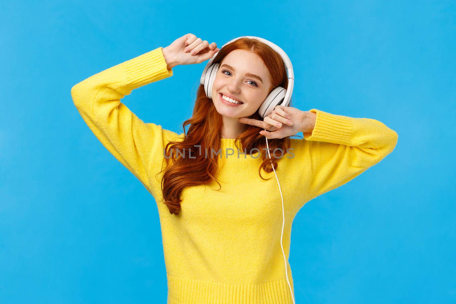 Technology, e-shopping and people concept. Modern good-looking redhead female in yellow sweater, dancing carefree, listen favorite singer music in headphones, smiling camera, blue background.