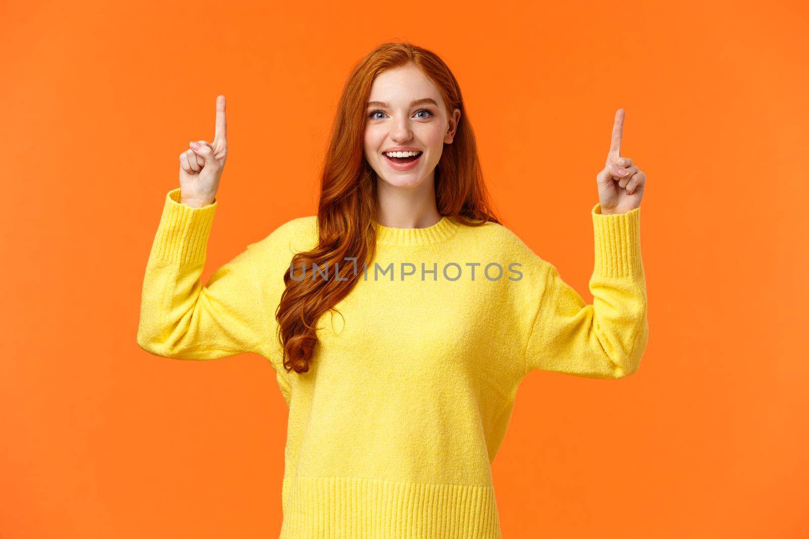 Excited, tender good-looking redhead girl with freckles, wear yellow sweater, pointing up and smiling joyfully, telling about promotion, advertising top product, orange background.