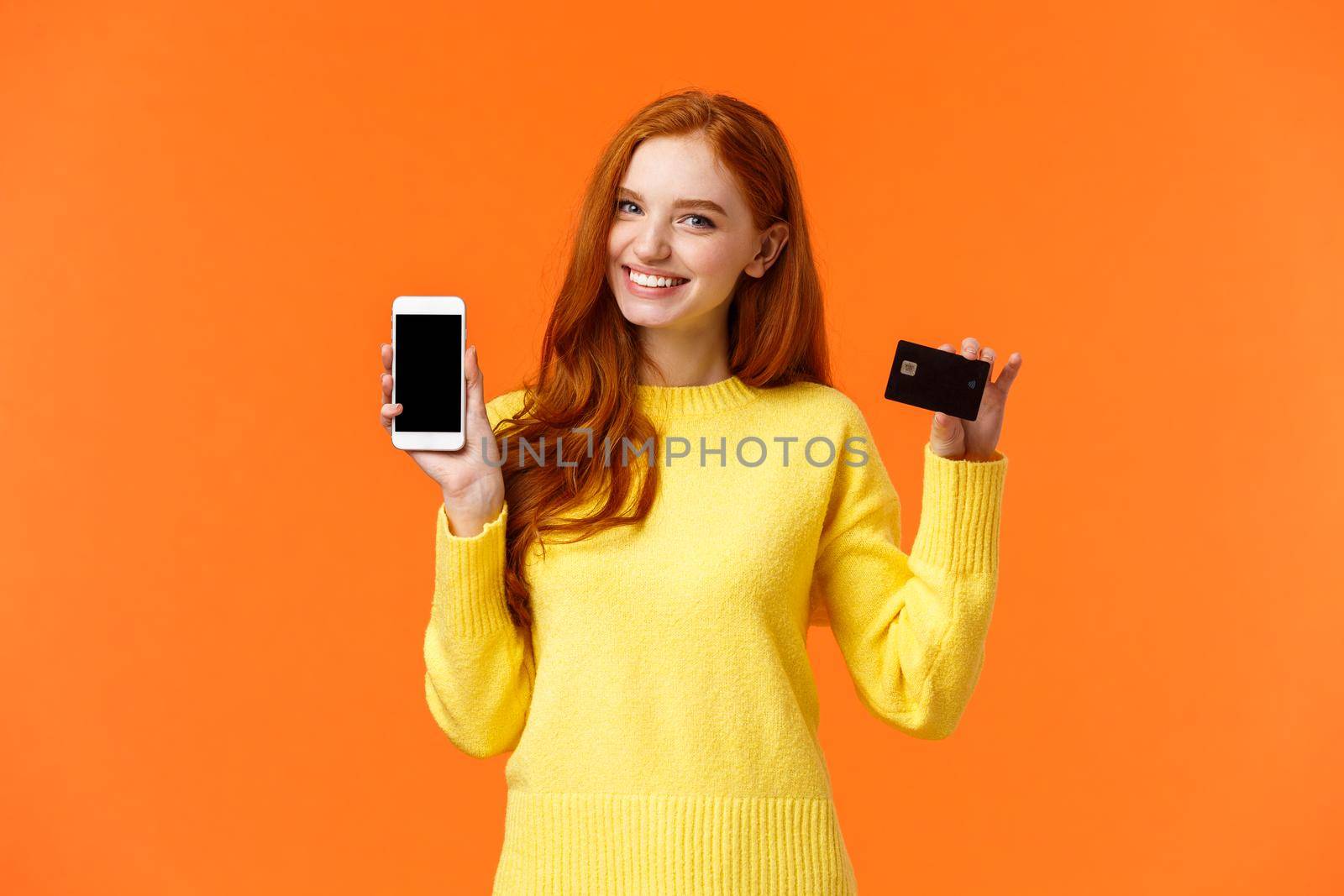 Redhead caucasian girl showing application on smartphone as holding mobile phone and credit card, using mobile banking app, cheerfully smiling camera, advertising over orange background.
