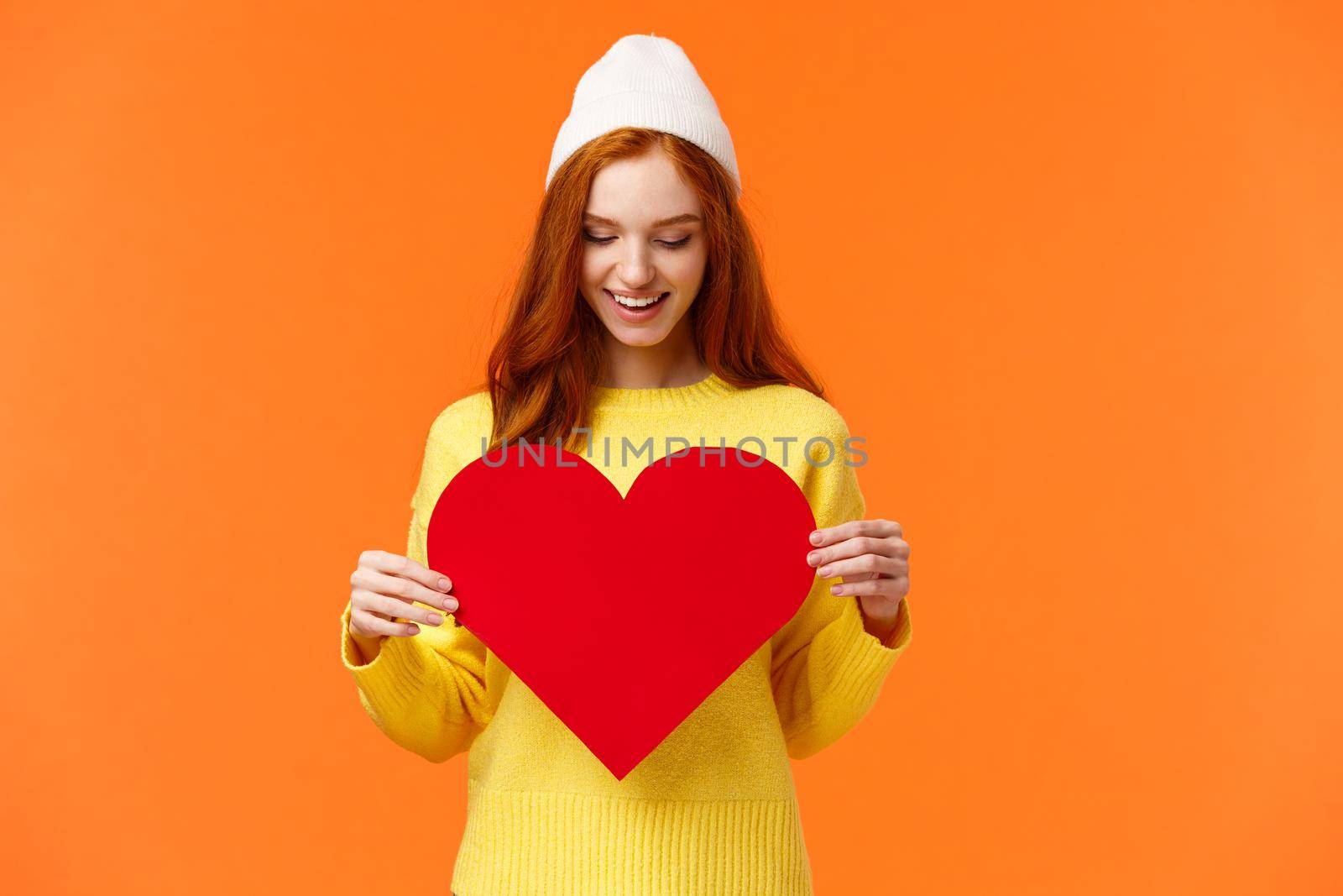 Waist-up portrait lovely romantic girl prepare gift for girlfriend on valentines day, asking be my valentine holding big red heart sign and smiling silly, orange background, wear winter hat.