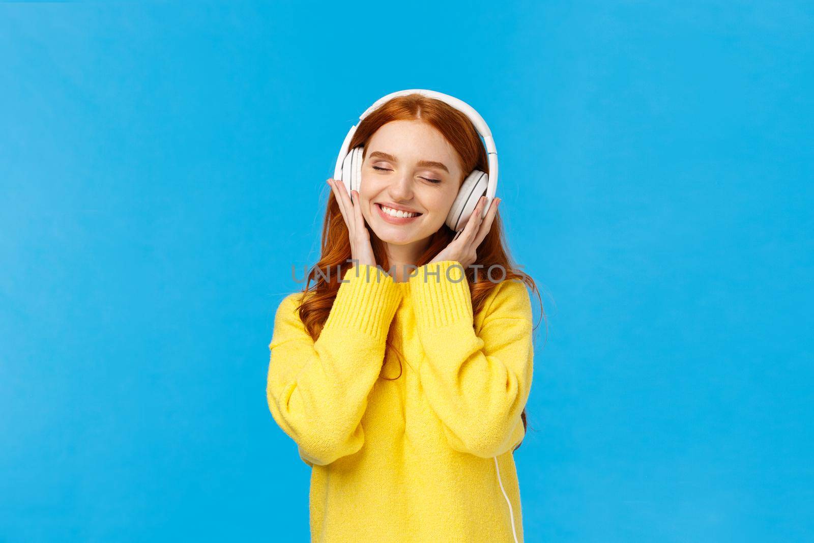 Technology, gadgets and lifestyle concept. Charming carefree redhead woman enjoy listening music in large white headphones, close eyes and smiling romantically, standing blue background.