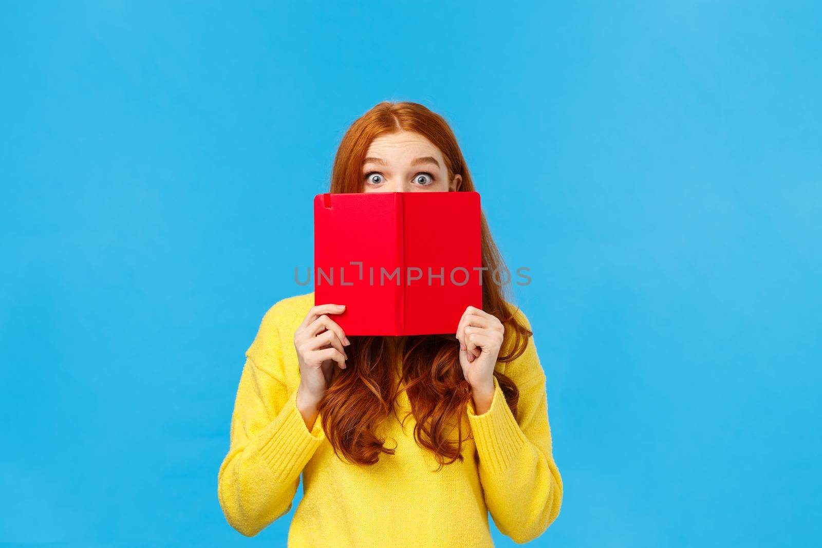 Shy and cute redhead girl like reading books and writing in diarty, hiding face behind red cool notebook, stare curious and surprised with popped eyes, standing blue background modest.