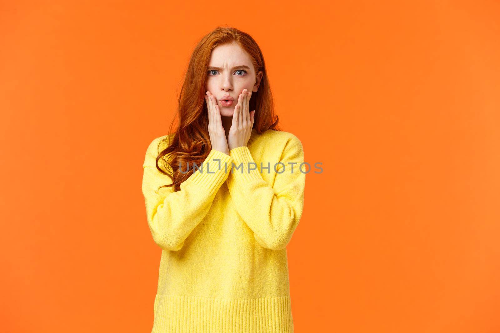 Woman looking surprised and startled with news. Astounded young redhead girlfriend found out secret, look tensed and shocked, gasping, folding lips, frowning serious, touch face, orange background.