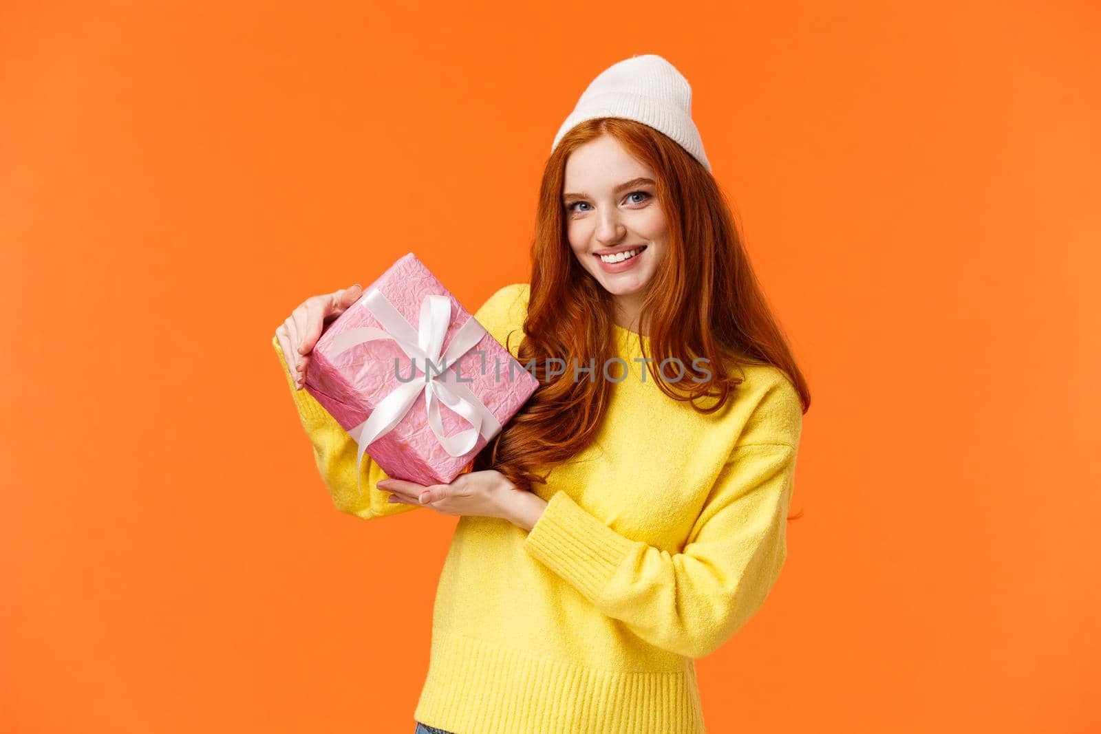 Lovely and romantic cute redhead woman prepared gift for valentines day, wrapped present in pink paper, showing box friend and smiling cheerful, standing upbeat orange background in winter hat.