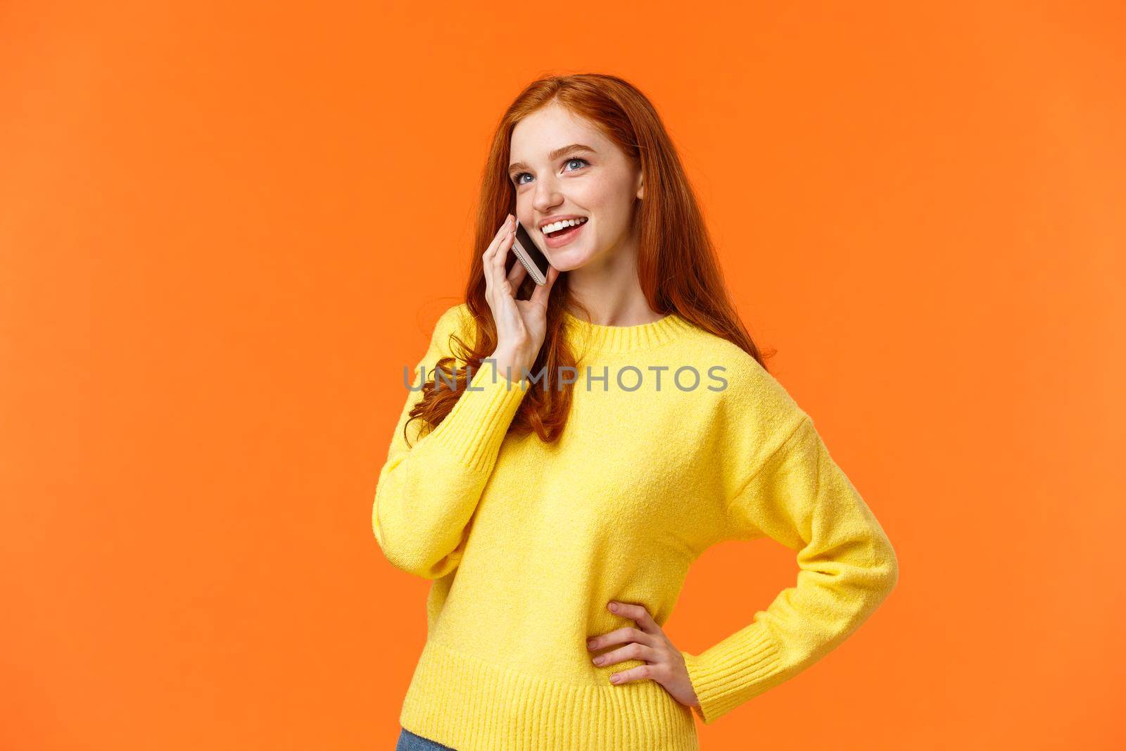 Outgoing pretty redhead girl having conversation on phone, calling friend, making arrangements, woman booking hotel or trip using mobile, speaking via smartphone, standing orange background.