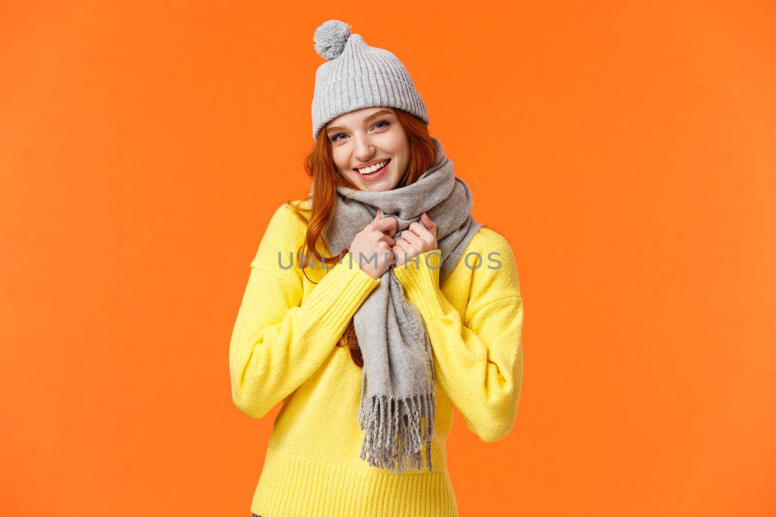 Its getting cold outside. Cute and lovely pretty redhead female wear grey winter hat and scarf, getting warm prepare play snowballs with friends outside during winter holidays, orange background.