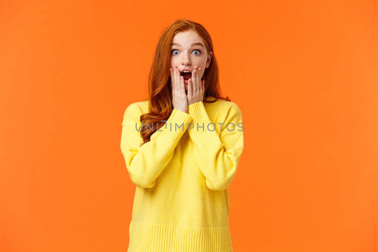 Impressed and speechless redhead woman found out awesome news, gasping, drop jaw astonished, touch cheeks and stare startled camera, christmas shopping season started, orange background.