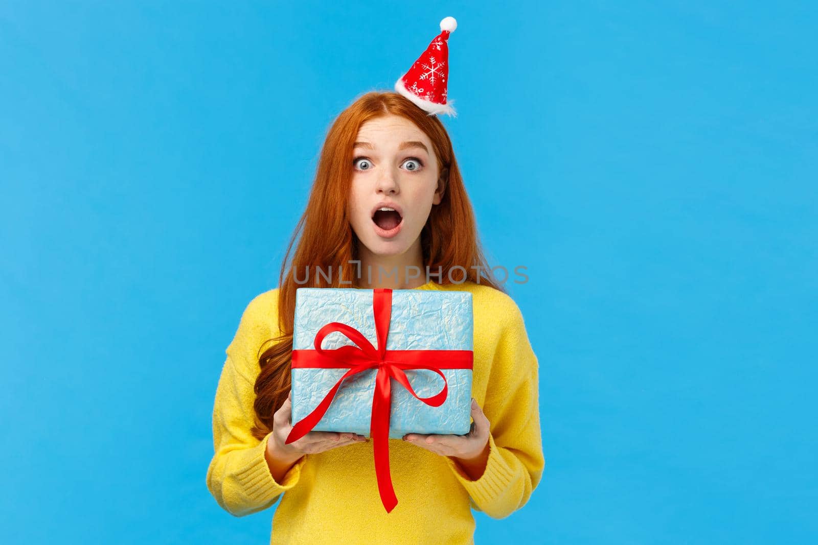 Surprised startled pretty redhead girl receive unexpected gift for christmas classmate, open mouth astonished and speechless staring camera as holding cute blue present wrapped box, studio background.