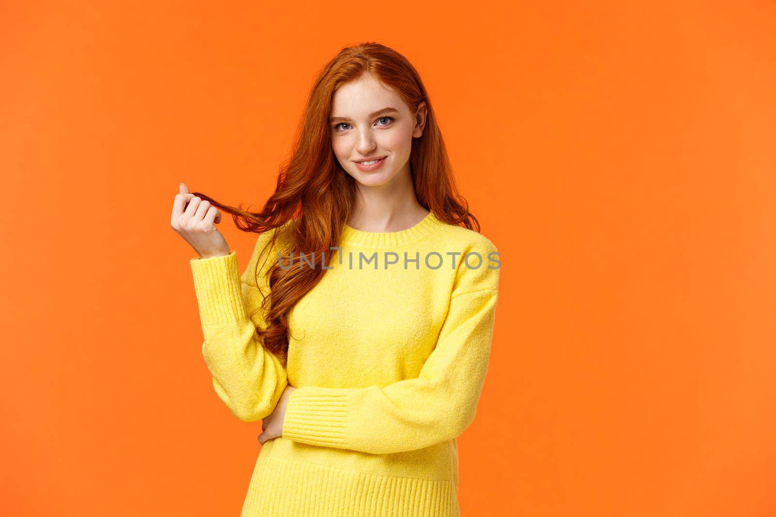 Gorgeous flirty and cute young modern redhead cheeky girl, rolling curl on hair and glancing camera curious, with interest and coquettish flirting smile, standing orange background intrigued.