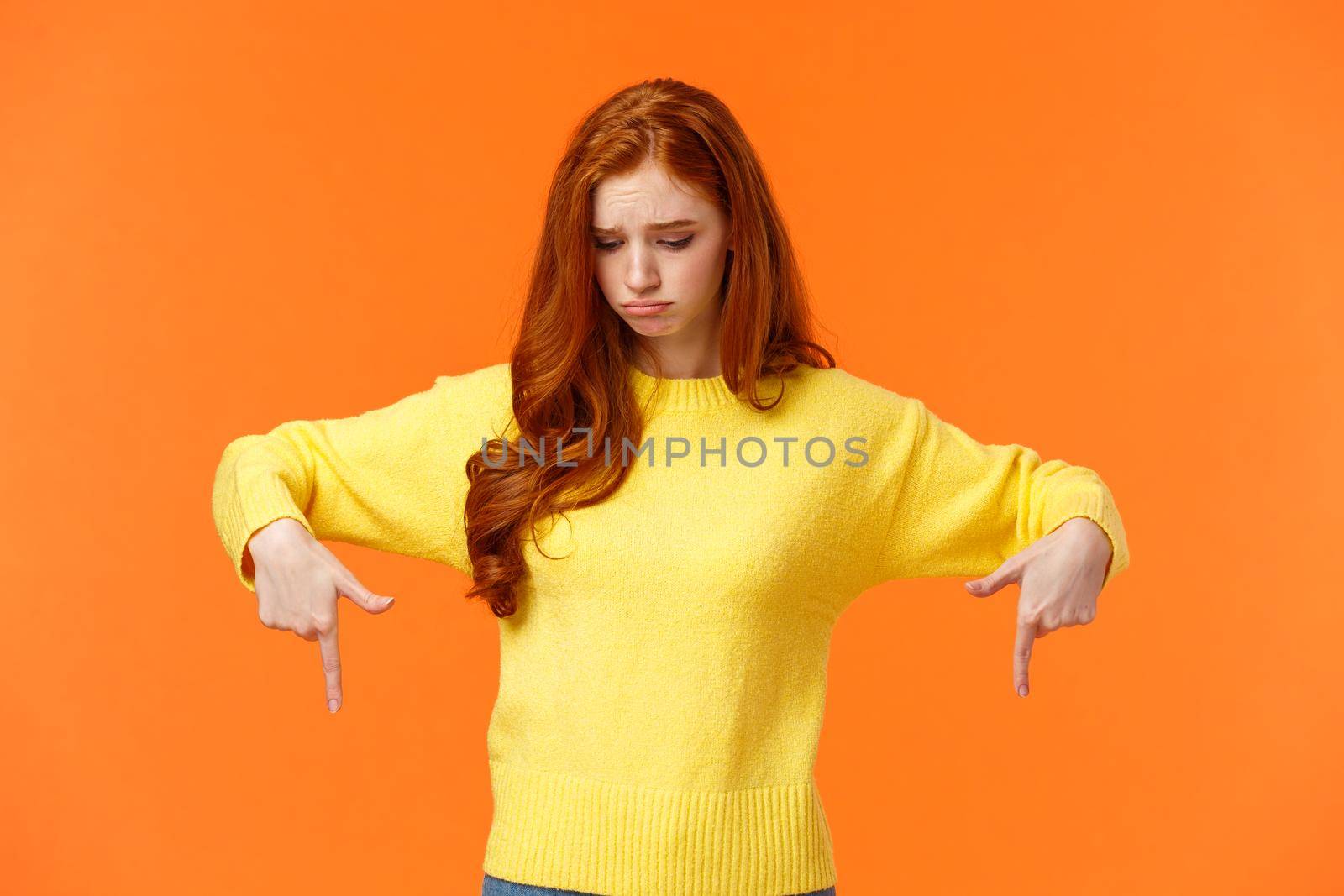 Envy, sad and sulky cute redhead woman in yellow sweater, looking and pointing down with jealousy or disappointment, feeling jealousy and regret missing chance, standing orange background.