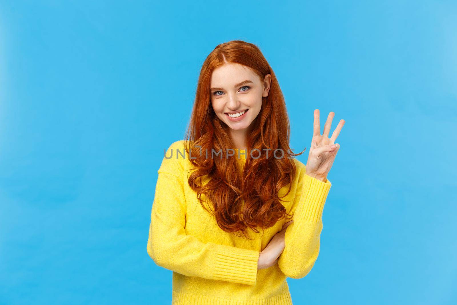 Gorgeous young redhead girl 20s in yellow sweater, smiling carefree making order, reservetion for three people, showing number third with fingers, standing blue background by Benzoix