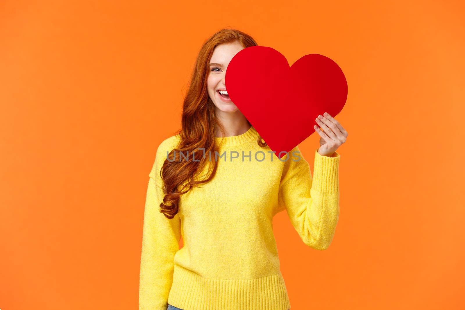 Holidays, romance concept. Cheerful girl showing affection, give heart to girlfriend, hiding half face behind valentines card, smiling joyfully, prepare romantic gesture for date, orange background by Benzoix