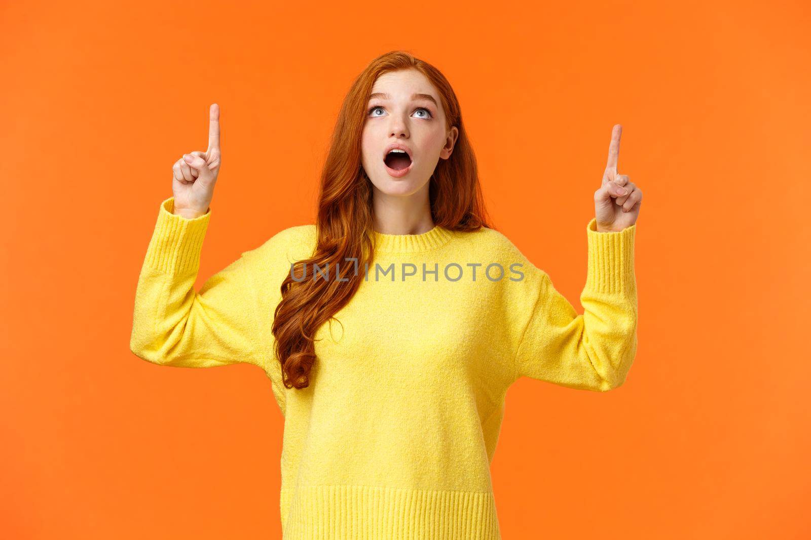 Wow amazing. Astonished and surprised redhead girl checking-out holiday special promo, woman stare and pointing up, stumble uppon cool offer, standing orange backgound astounded.