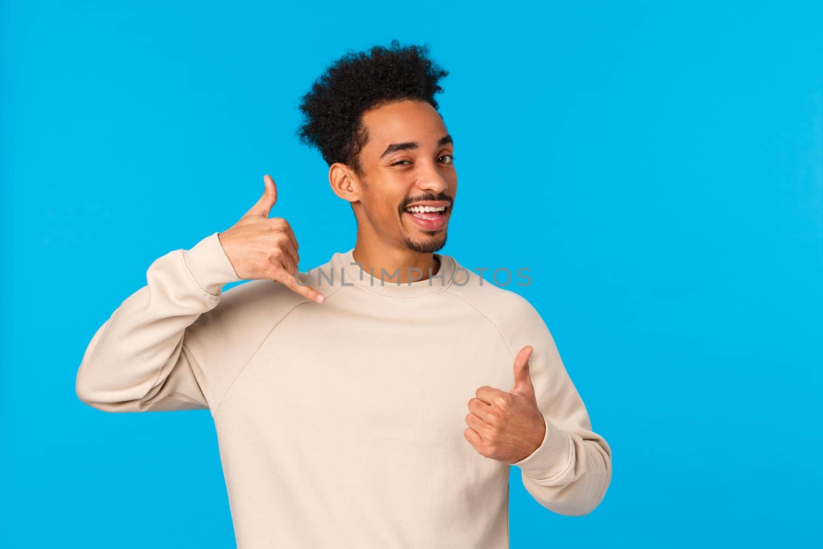 Call me later babe. Sassy and coquettish african-american confident macho guy with moustache, afro haircut showing thumb-up and phone gesture, wink and smiling flirty, blue background by Benzoix