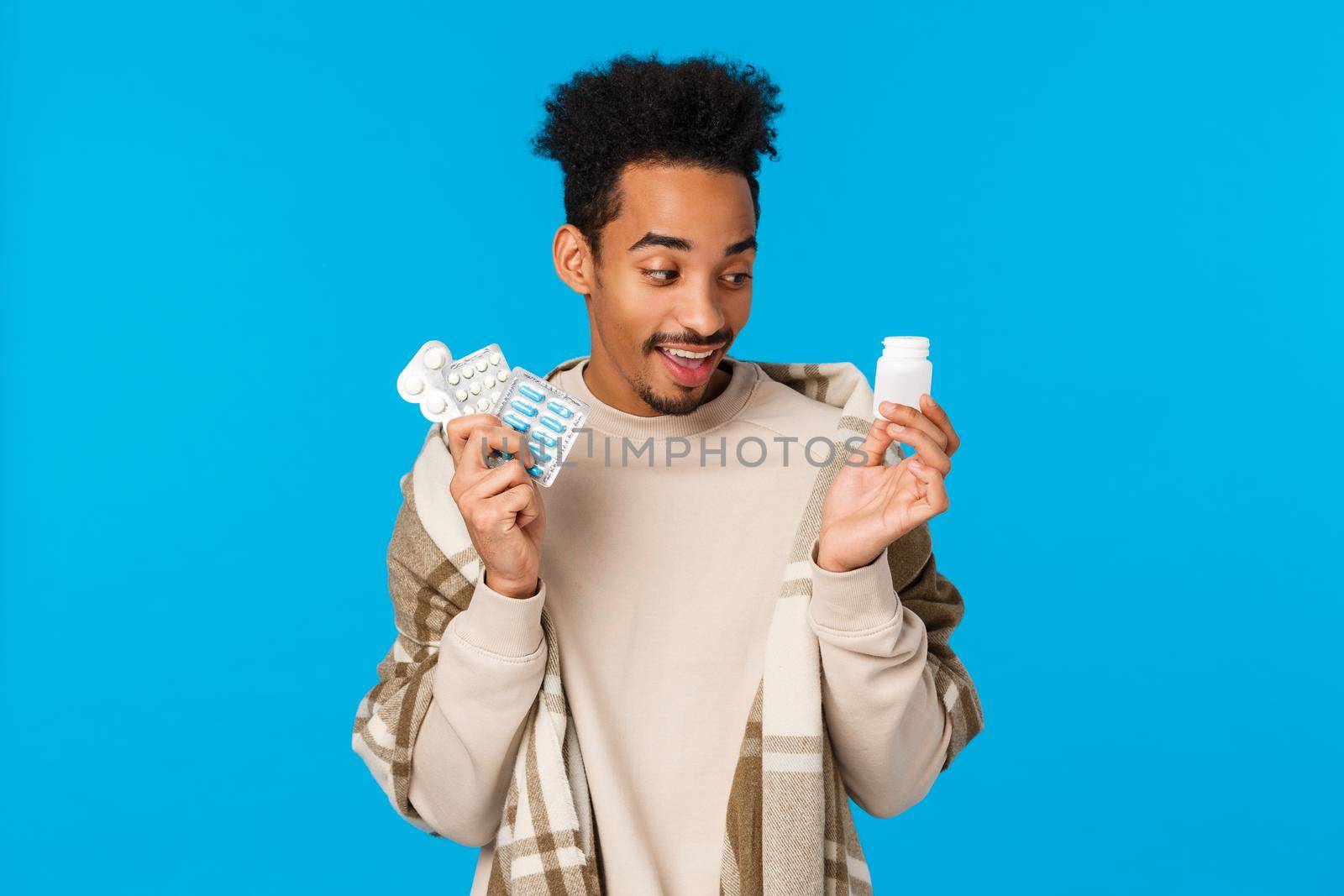 Funny handsome african man choosing what pill take first as got sick, staying home with cold or flu, holding pills from drugstore, use medication get better, taking care health, blue background.