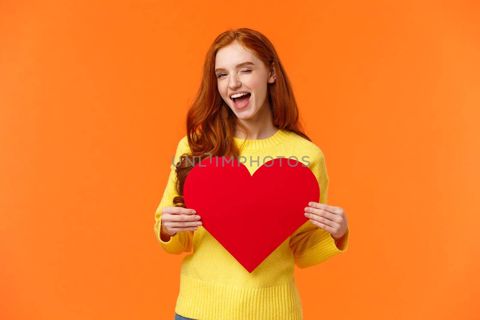 Hey gorgeous you have my heart. Valentines day, boyfriend and girlfriend concept. Cute and sassy redhead woman holding large sign and smiling, wink coquettish, standing orange background.