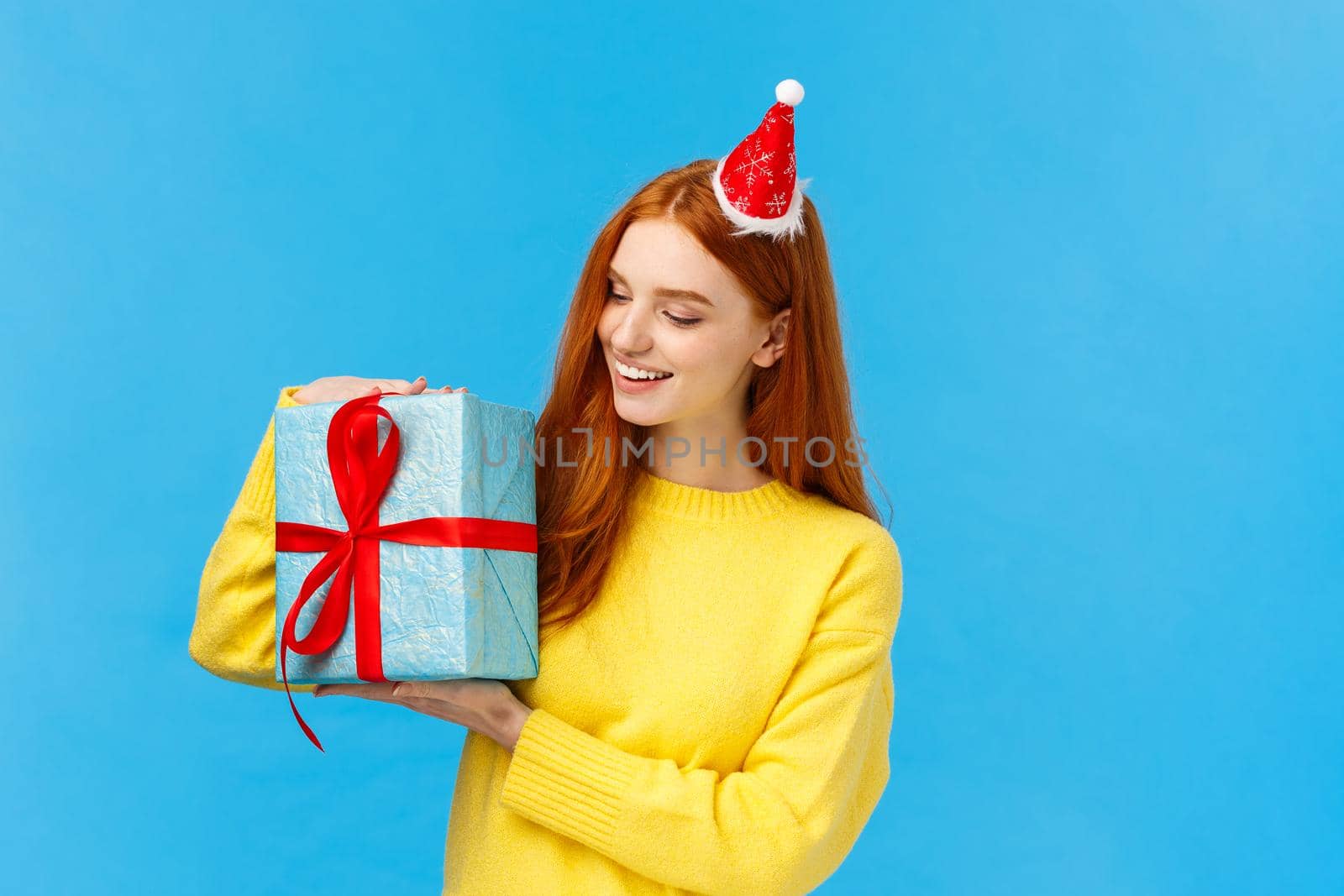 Christmas, new year and holidays concept. Attractive lovely redhead woman, girlfriend in cute hat holding wrapped gift, curious whats inside, smiling intrigued looking at present, blue background.