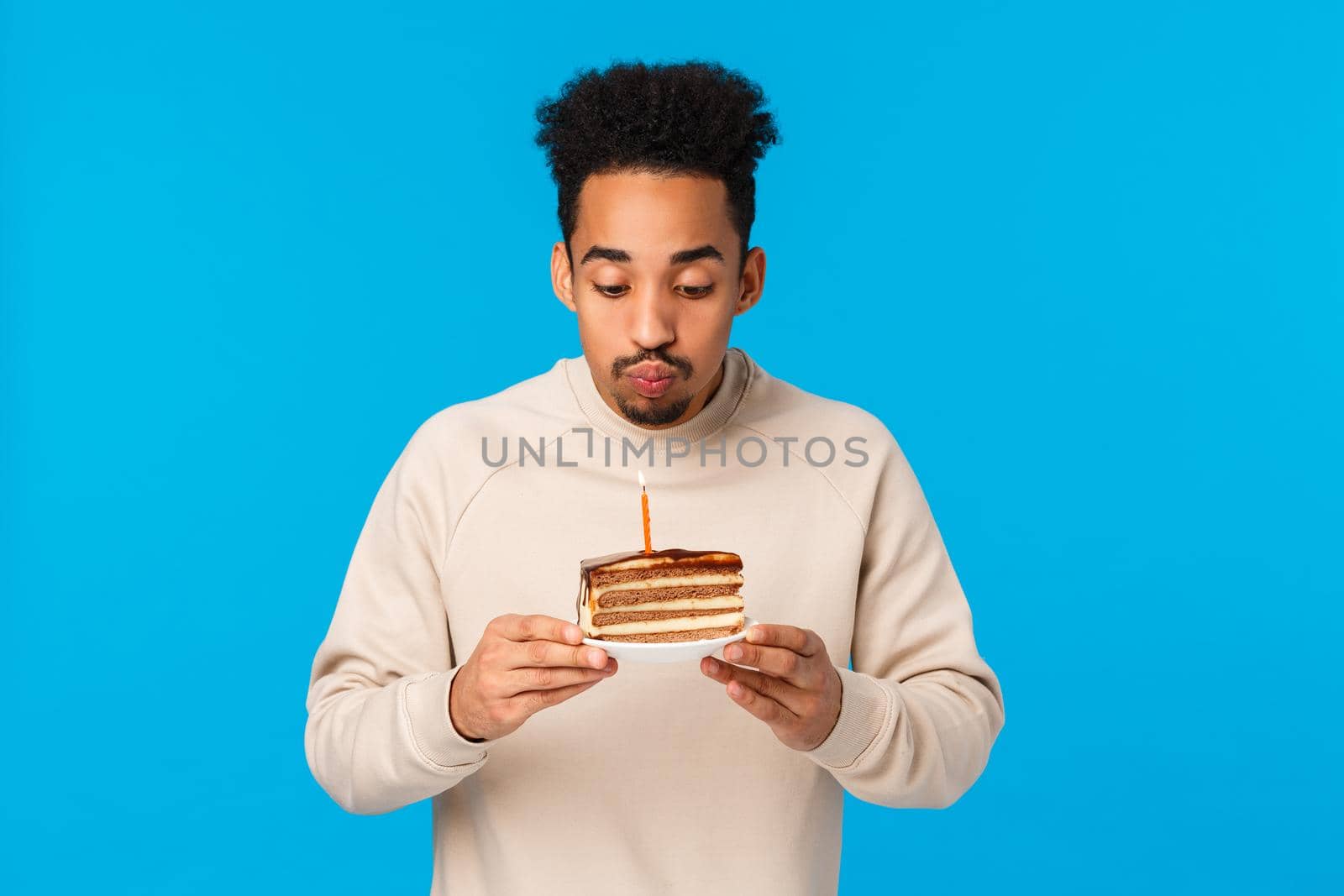 Celebration, party and happiness concept. Waist-up portrait african american joyful guy pouting, blowing-out candle on b-day cake, enjoying birthday making wish, standing blue background.