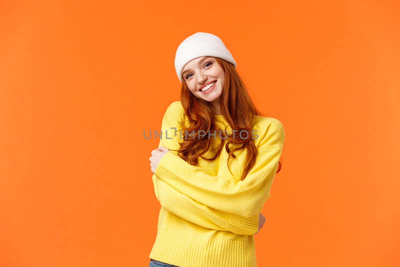 Winter is her favorite season. Tender silly and lovely redhead girl embrace herself and tilt head, smiling cute, wearing beanie and soft warm sweater just for cold weather, orange background by Benzoix