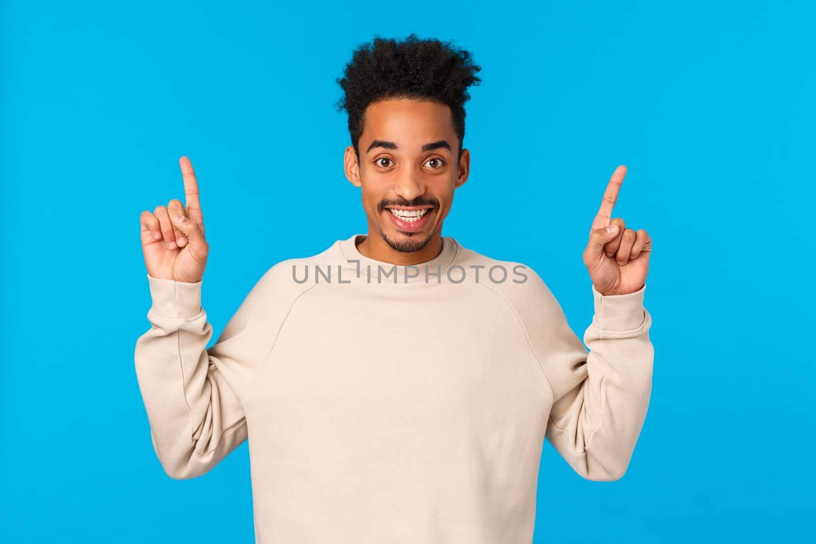 Cheerful handsome african-american young hipster guy with moustache, afro pointing fingers up, telling about promo, looking camera satisfied and beaming smile, standing blue background by Benzoix