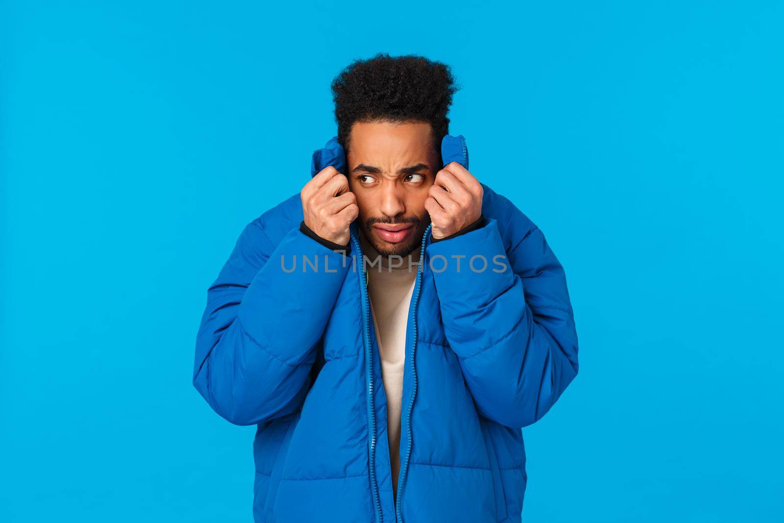 Embarrassed or ashamed guy hiding his face not to be recognized in public. Nervous and alarmed african american man pull collar of padded jacket on face and peeking left, avoiding person by Benzoix