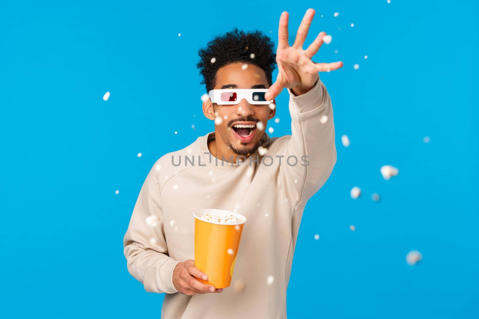 So realistic I want touch it. Entertained and amused happy african-american hipster guy in movie, wearing 3d glasses extend hand and throwing popcron to grab animated thing, blue background.