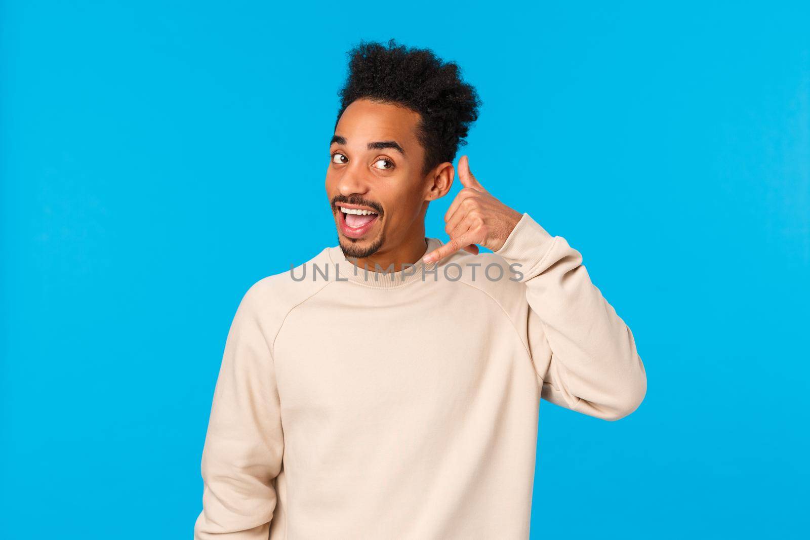 Hello whos calling. Funny attractive and cheerful hipster african american guy with afro haircut, moustache, imitating phone-call making mobile gesture near ear and answer, blue background by Benzoix