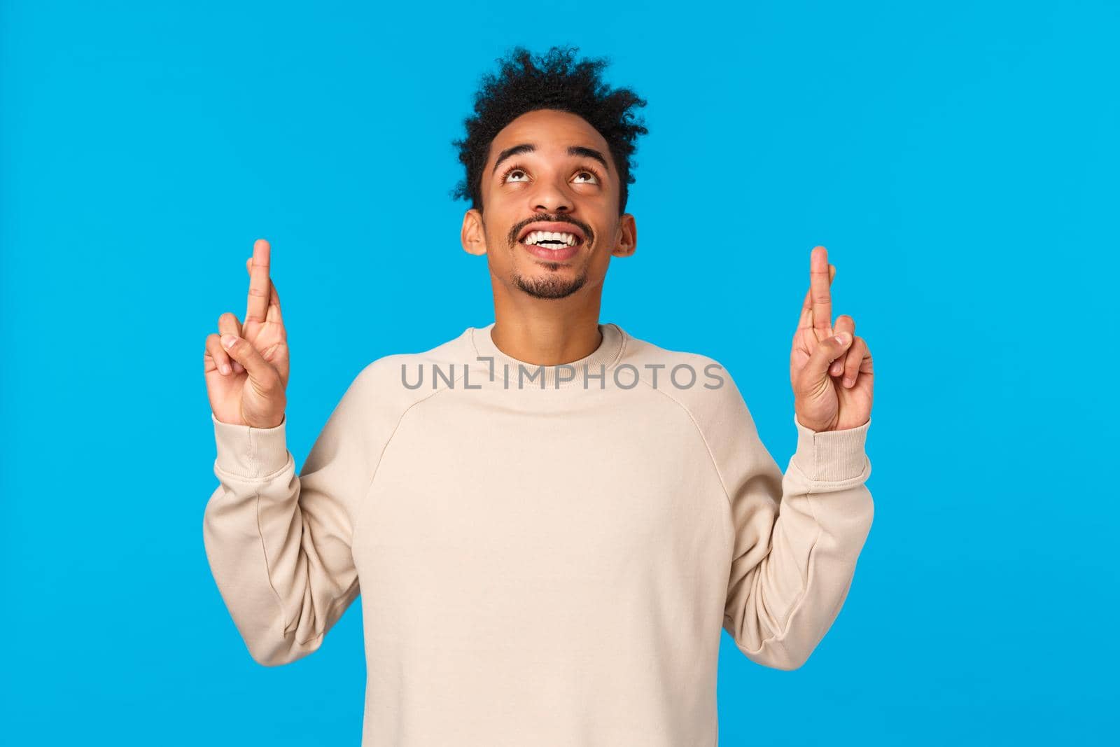 Enthusiastic and intrigued modern african-american male in white sweater, seeing something curious upwards, look and pointing fingers up, smiling astonished, wonder whats there, blue background.