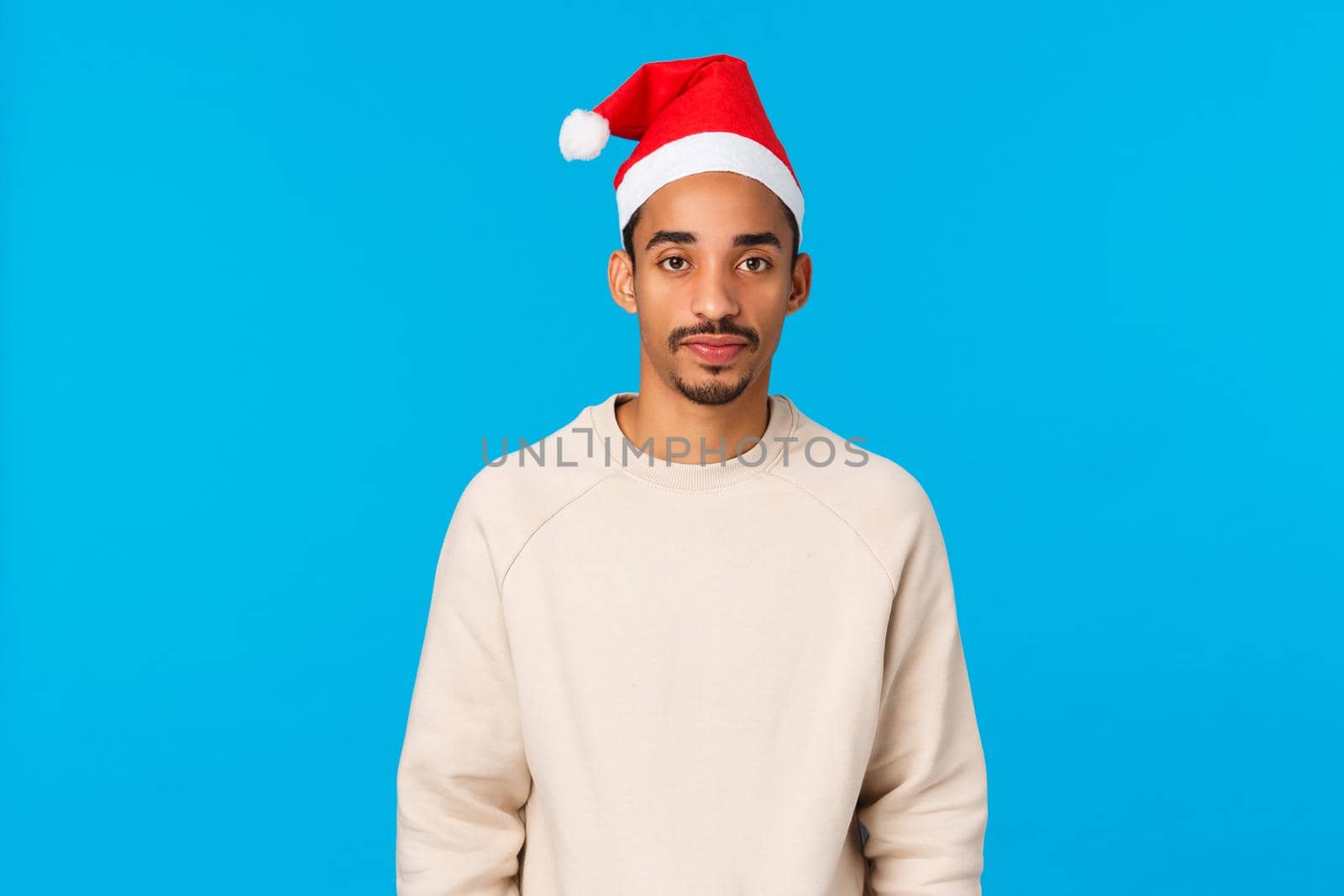 Christmas, holidays and family concept. Cute and silly african-american guy celebrate winter holidays parents house, wearing santa hat feeling awkward, attend new year party, blue background.