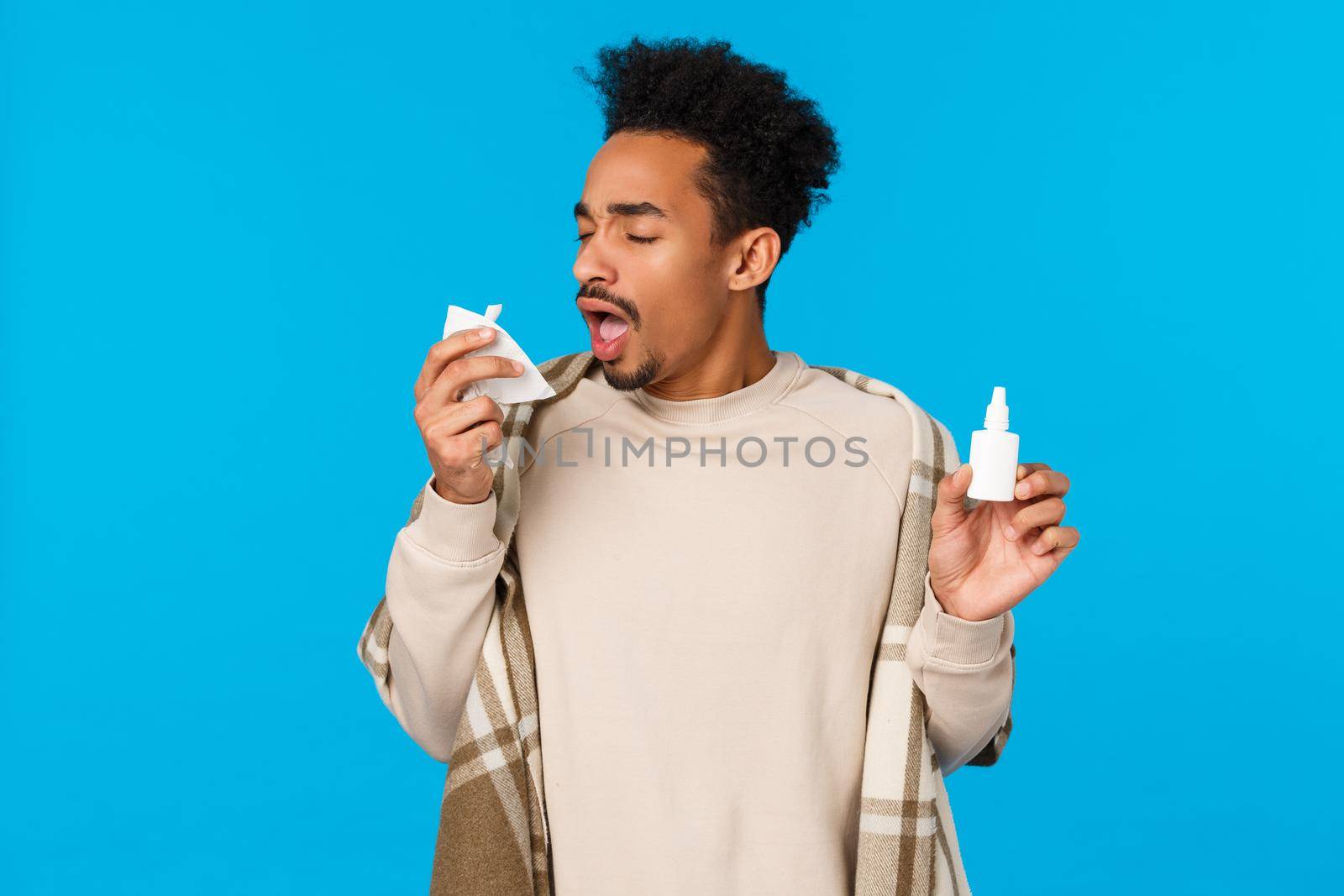 Ahchoo. Sick african american guy staying at home, skip college feeling unwell, caught cold, sneezing in napkin and holding nose spray bottle, use prescribed medication get better, blue background.