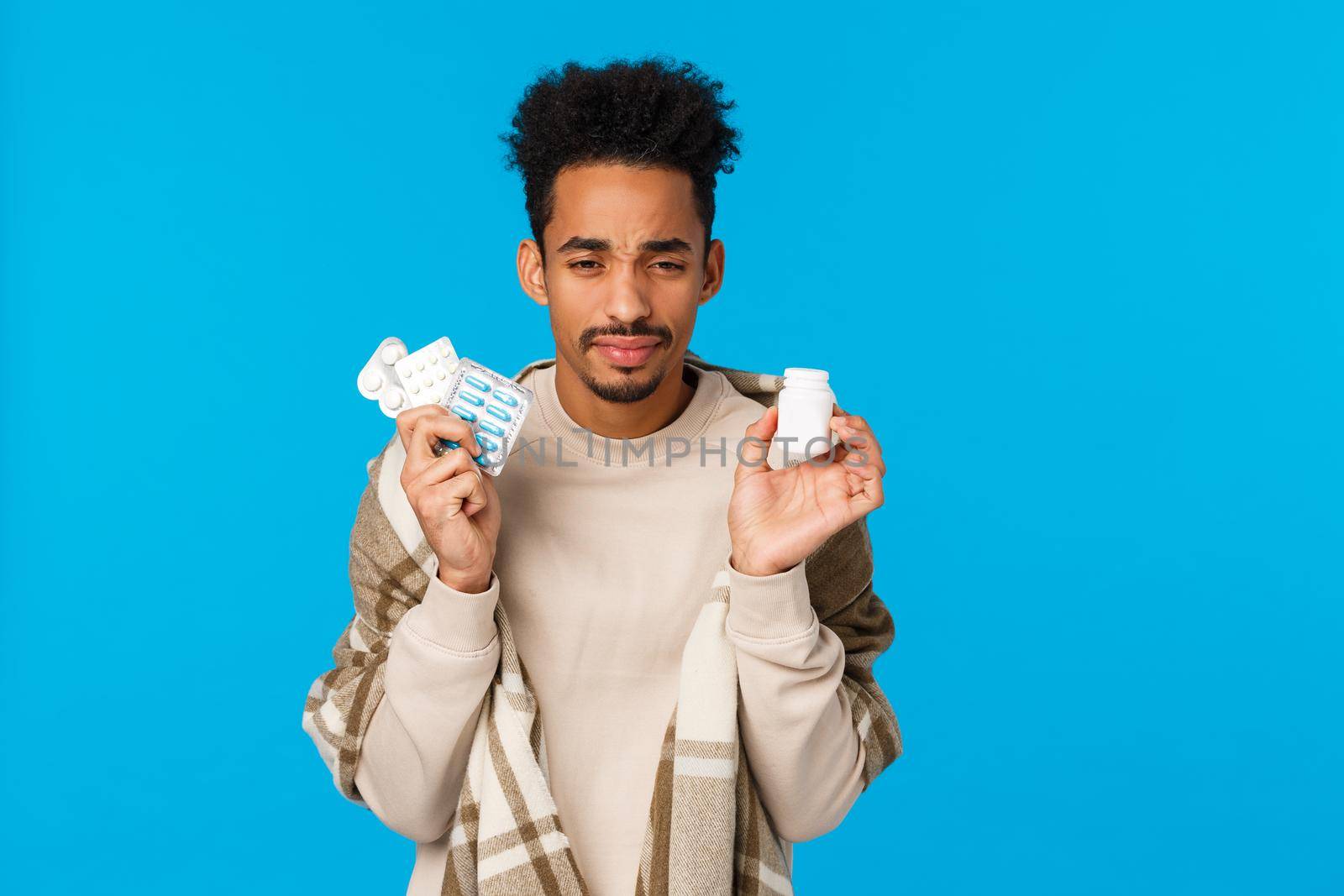 Health, medication and winter season concept. Young african american guy holding drugs and bottle of pills, sneezing, feel unwell fatigue from flu, caught cold, standing in blanket blue background by Benzoix