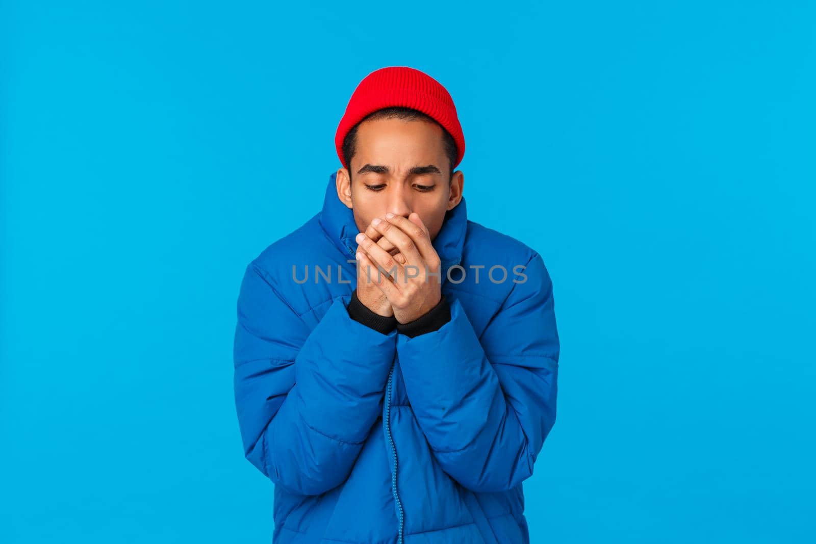 Man feeling cold, trying warm himself as blowing hot breathing at palms, trembling standing in snowy low temprature weather in blue padded jacket, red beanie, hate winter season. Copy space