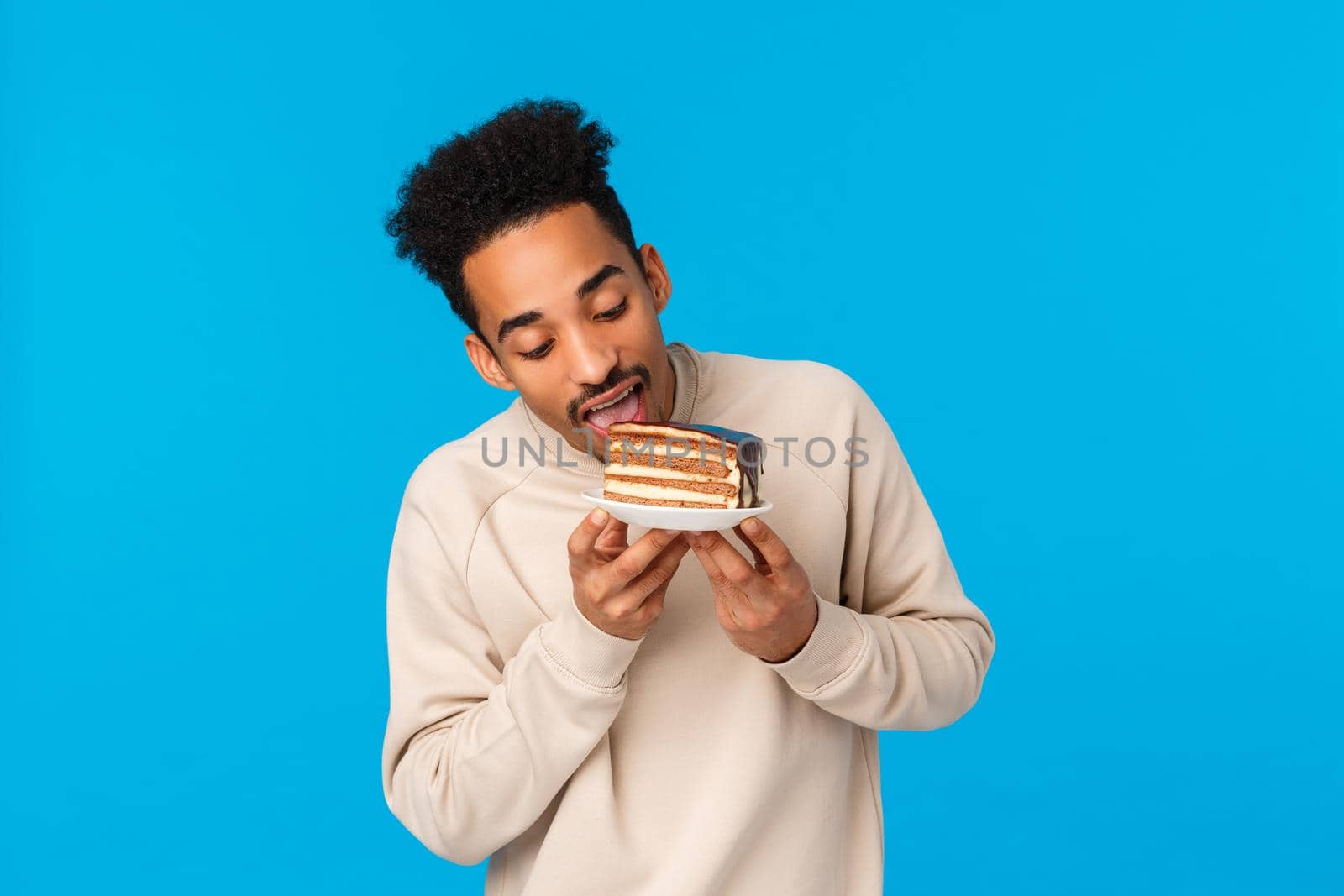 Happy holidays, sweets and tasty food concept. Handsome young african american stylish guy in sweater, taking bite of delicious chocolate cake, holding plate, skip diet, standing blue background.