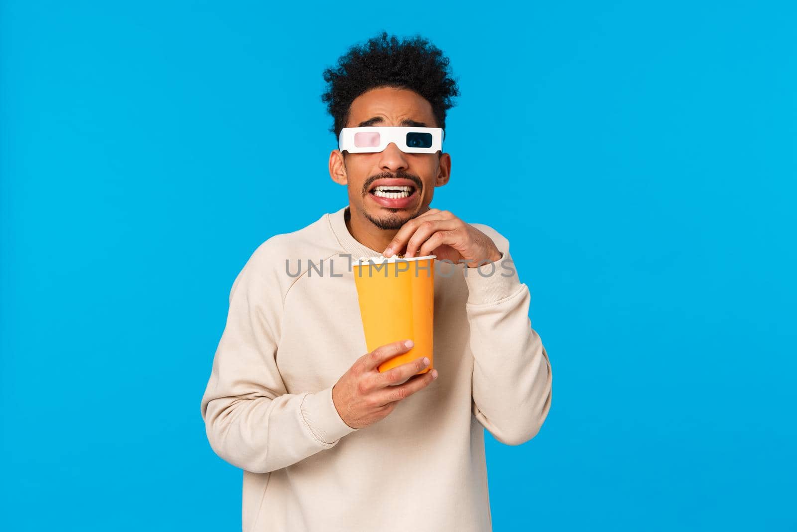 So scary. Trembling and terrified african-american hipster guy watching horror movie, frightening scene in film 3d glasses, holding paper box and eating popcron, standing tensed, blue background.