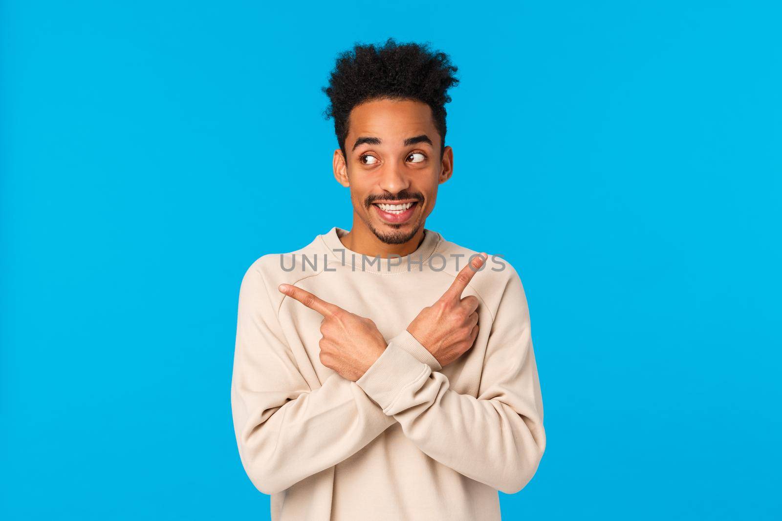 Guy making assumption what he get for holidays. Cute and excited charming african american hipster male in white sweater, pointing sideways left and right, smiling eager start celebrating, blue wall by Benzoix