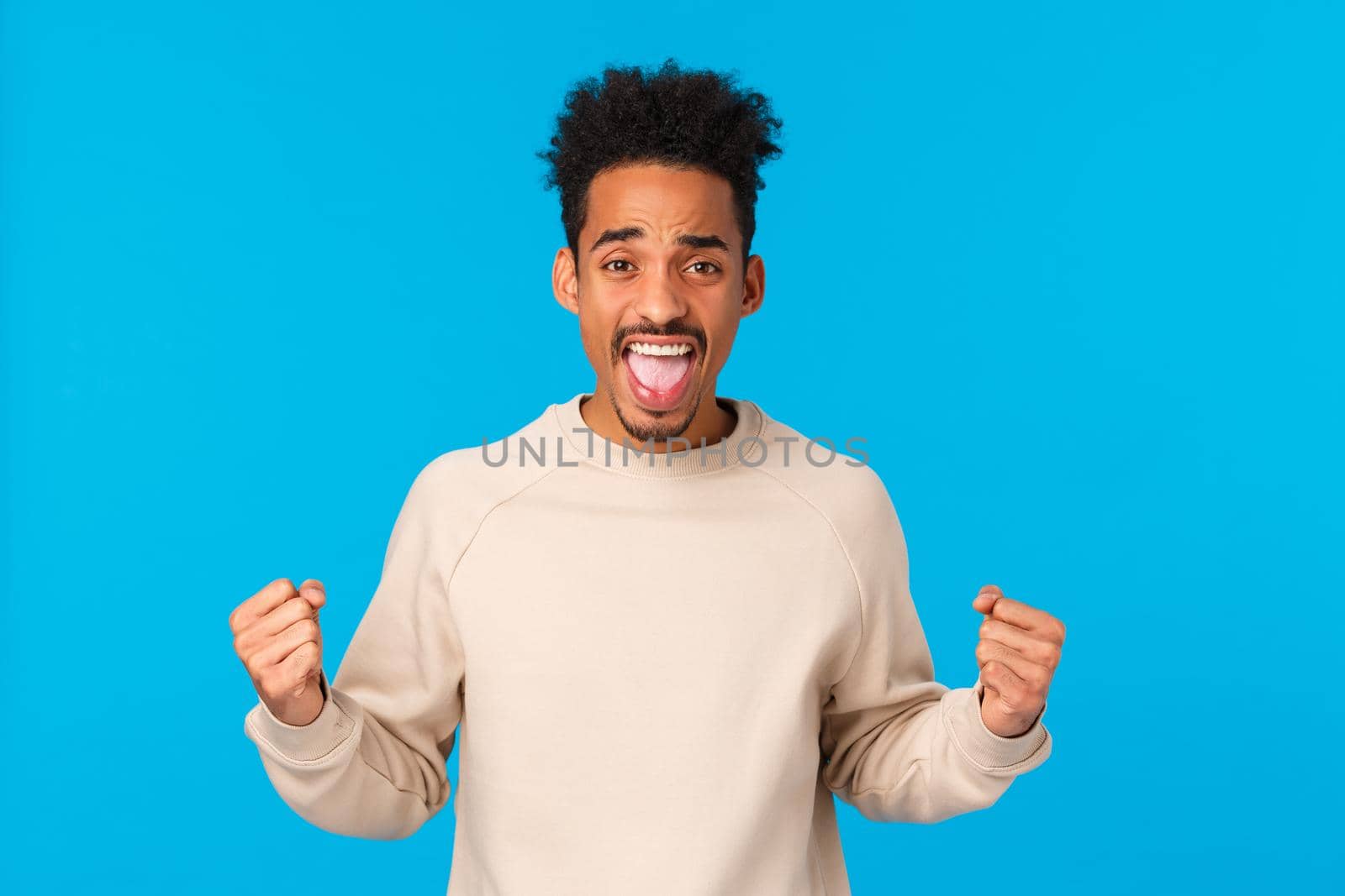 Cheerful and excited handsome african-american male with afro haircut, clench fists showing strength power, triumphing, rooting for team, watching sport game and cheering, blue background.