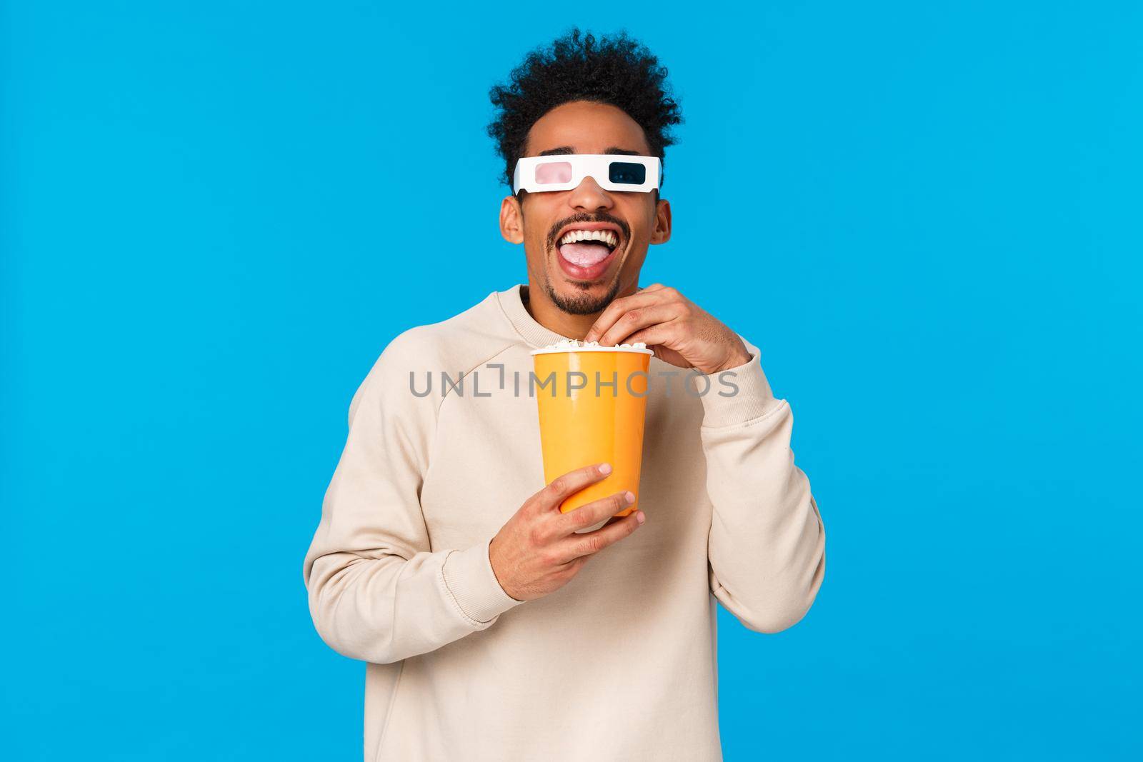 Entertainment, lifestyle and people concept. Carefree happy and relaxed african american man in white sweater, watching funny interesting movie, eating popcron and wearing 3d glasses, blue background.