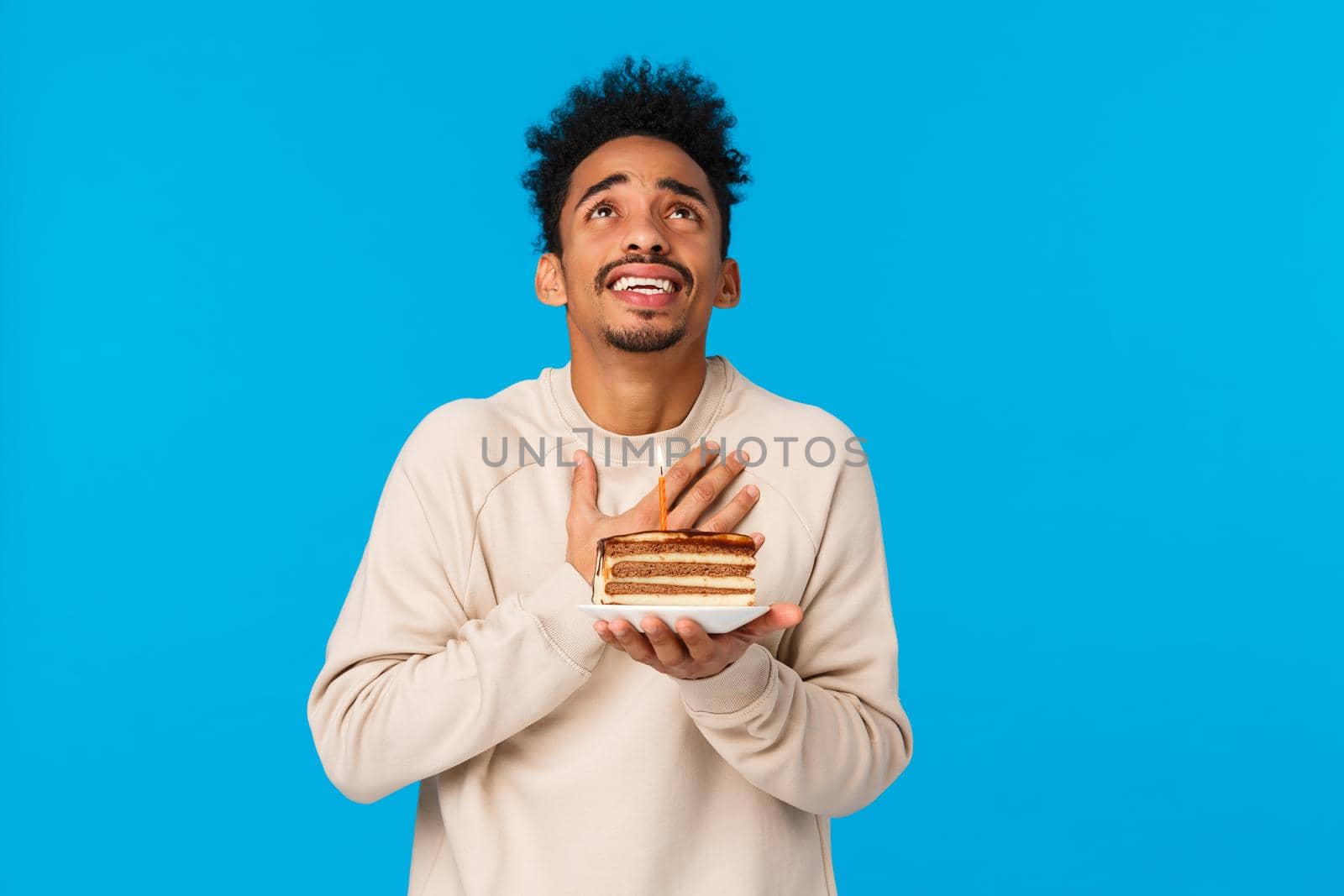 Sincere and hopeful african-american guy holding b-day cake with candle, holding hand on heart and looking sky as pleading make wish come true, feeling uneasy getting old on birthday, blue background by Benzoix