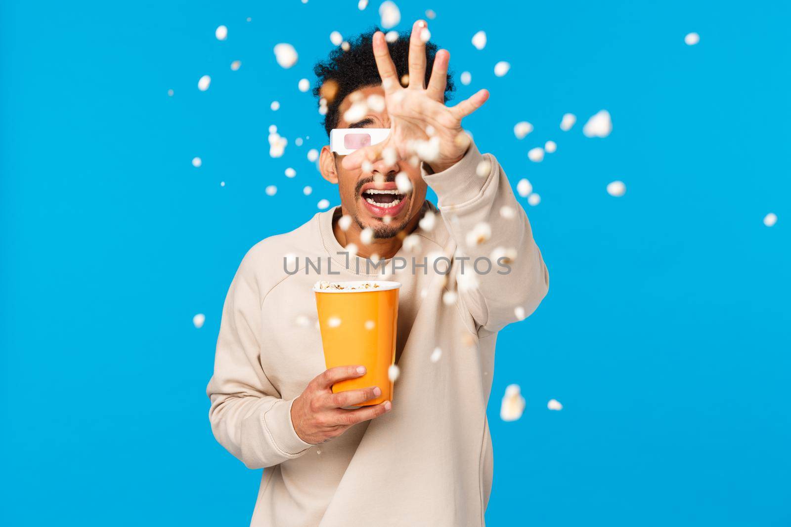 Scared and shocked young man watching 3d horror movie and throwing popcron at screen as feeling tensed and frightened from realistic picture, standing blue background afraid by Benzoix