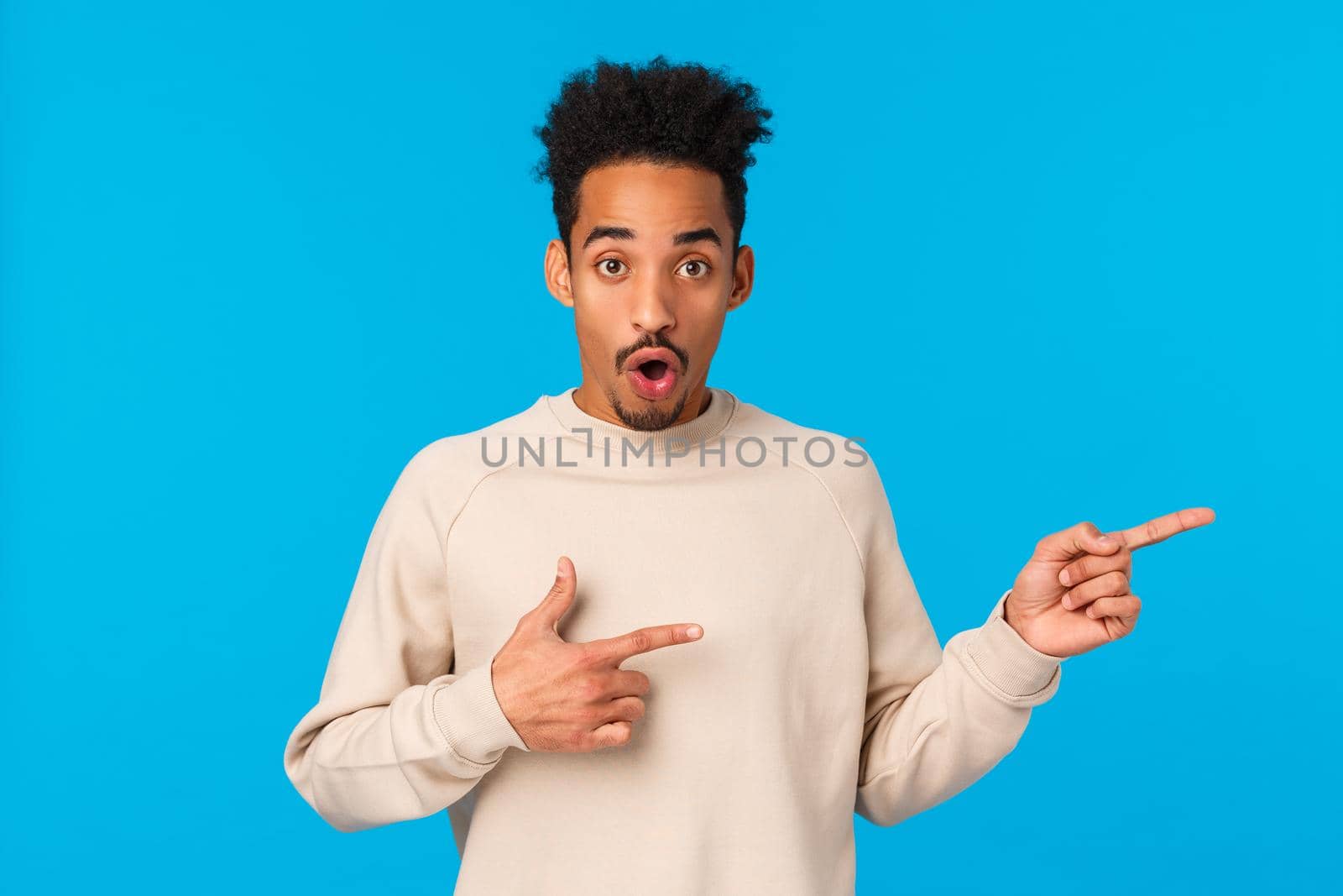 Questioned and impressed attractive african-american young male model with moustache, afro haircut, curious about event nearby, pointing fingers right, open mouth gasping amused, look wondered.