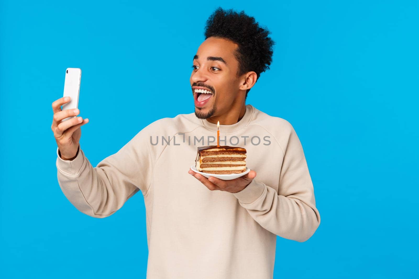 Excited happy african-american bearded hipster guy holding piece cake with birthday candle, smiling joyfully taking selfie or record video how he celebrates, making wish, standing blue background by Benzoix