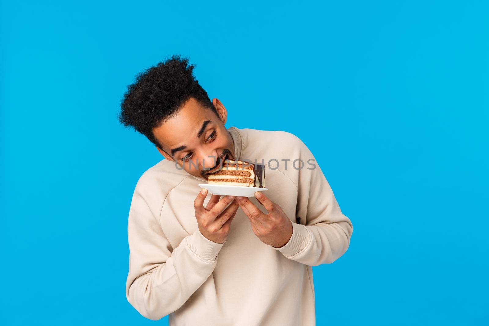 Guy stole piece cake and eating it fast. Funny and cute african-american man holding plate, bite delicious dessert celebrating birthday, enjoying holidays tasty food, standing blue background by Benzoix